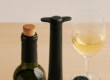 Een geopende fles wijn met een kurk erin, een zwarte vacuümpomp, een losse vacuümstopper en een ingeschonken glas witte wijn op een lichte houten ondergrond
